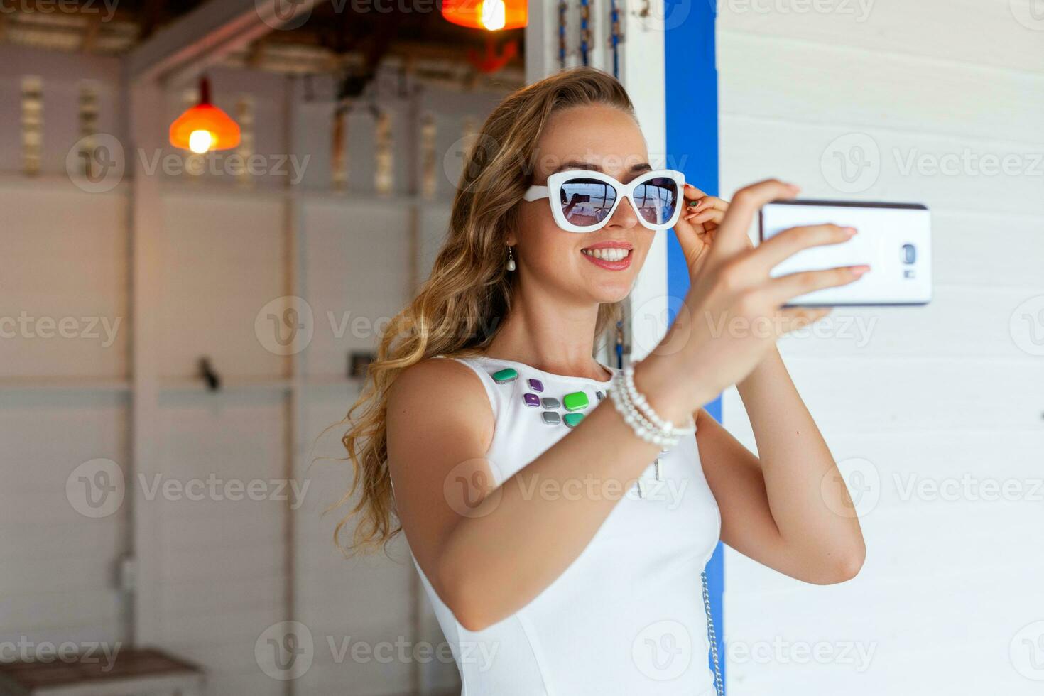 attractive woman in white dress in summer cafe in sunglasses using phone taking photo