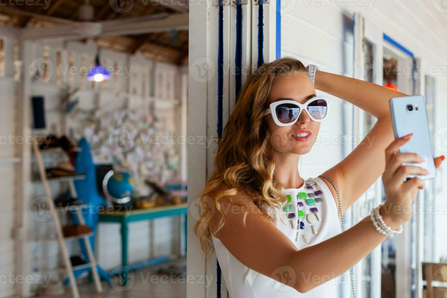 attractive woman in white dress in summer cafe in sunglasses using phone taking photo