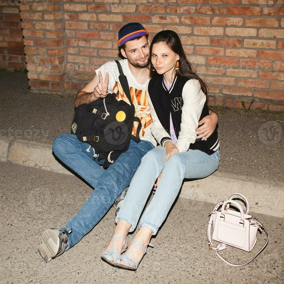 joven elegante hipster Pareja en amar, estilo atuendo, vaqueros, abarcar, frio accesorios, sentado en suelo en contra ladrillo muro, feliz, teniendo divertido foto