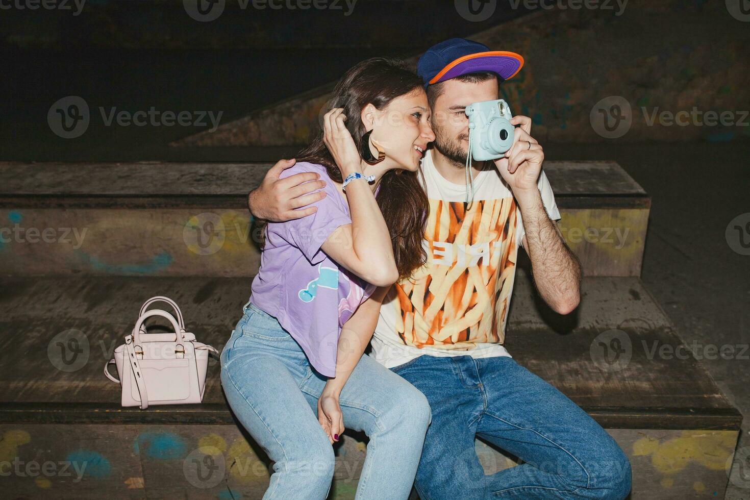 young stylish hipster couple in love, swag outfit, jeans, embrace, cool accessories, sitting happy, having fun, photo camera