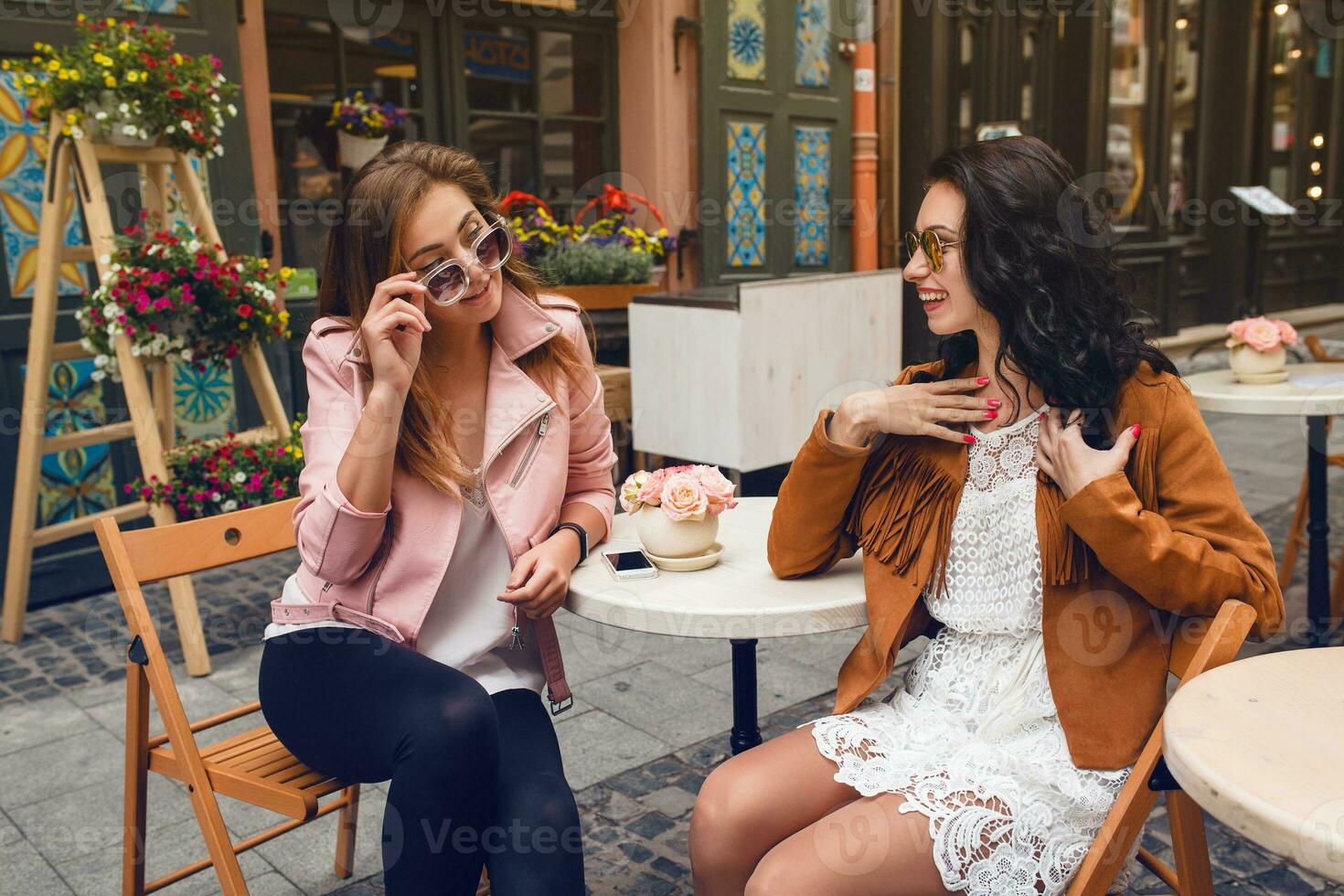 dos joven elegante mujer sentado a cafetería, hablando, chismoso, elegante de moda atuendo foto