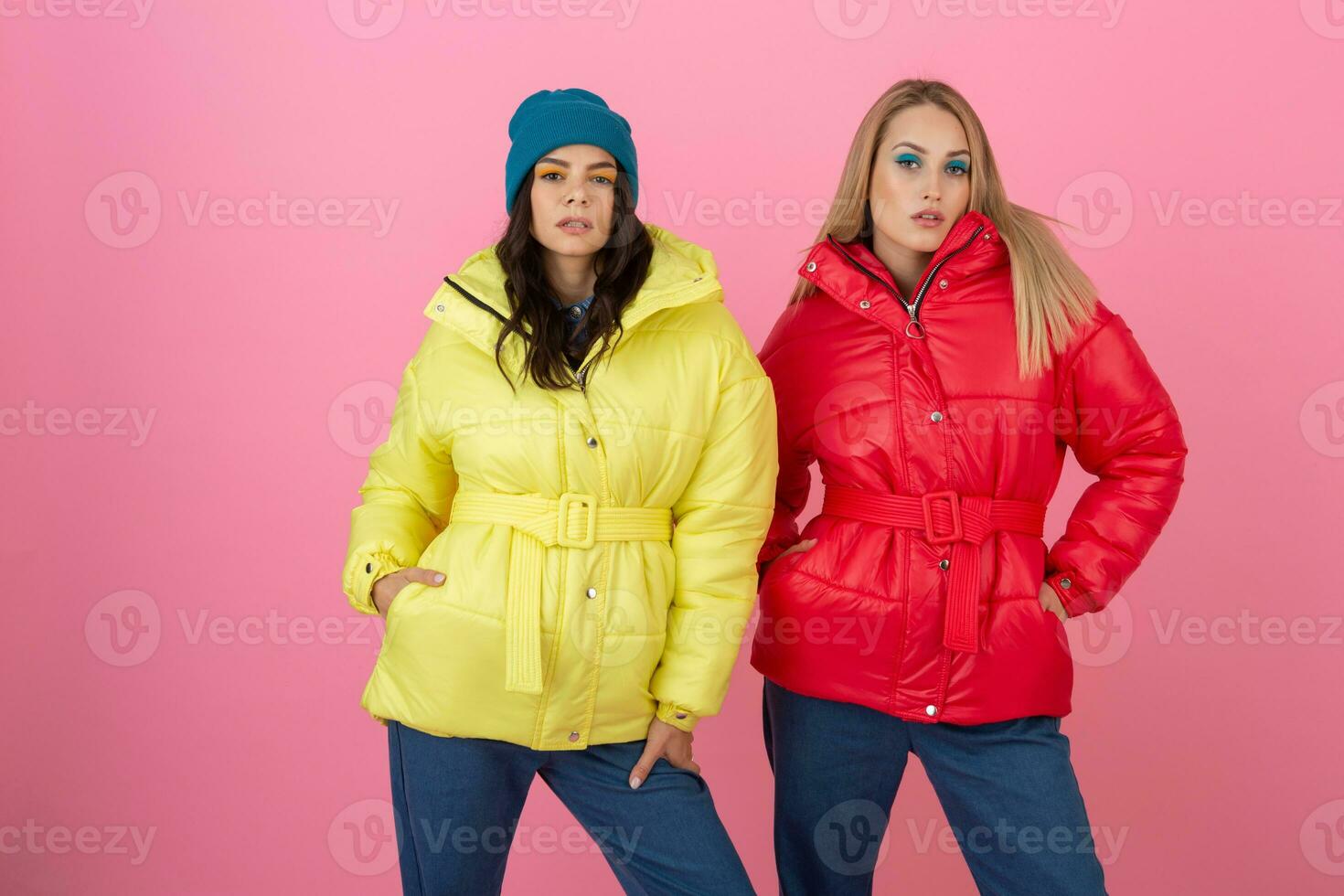 two attractive stylish women posing on pink background in colorful winter down jacket of red and yellow color, warm clothes fashion trend photo