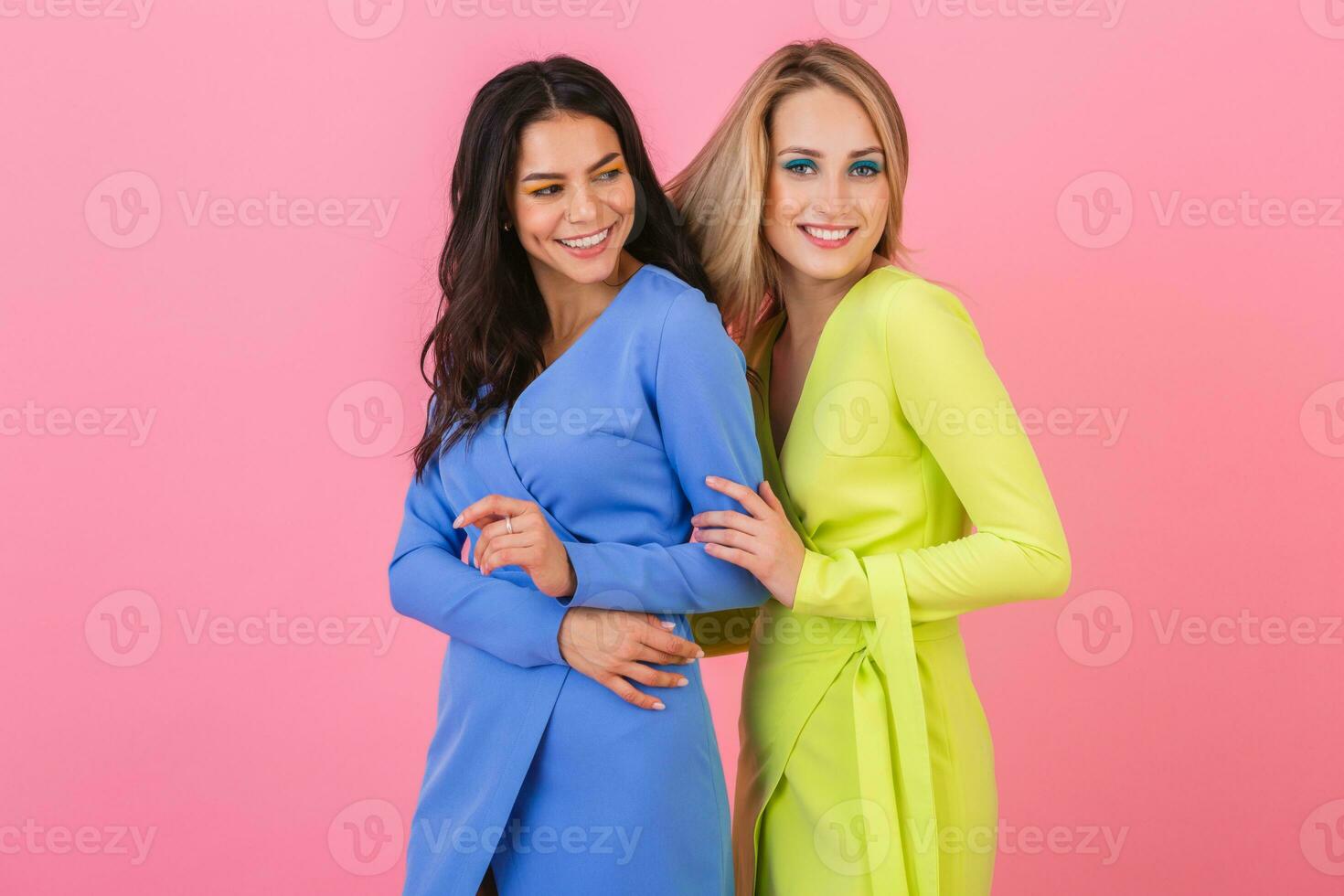 two stylish smiling attractive women friends posing on pink background in stylish colorful dresses of blue and yellow color, spring fashion trend photo