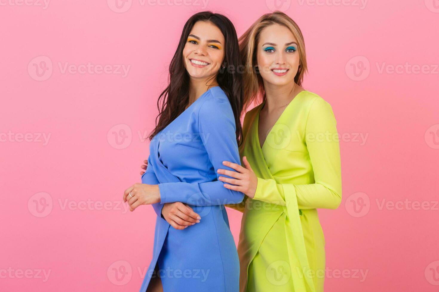 two stylish smiling attractive women wearing colorful dresses having fun posing on pink background, blue and yellow color apparel, summer fashion trend photo