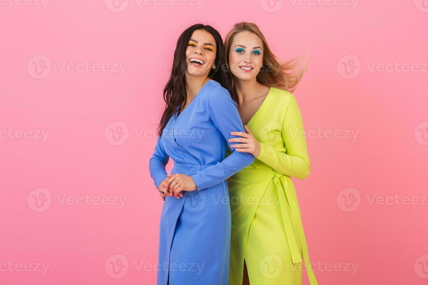 two stylish smiling attractive women having fun posing on pink background in stylish colorful dresses of blue and yellow color, spring fashion trend photo