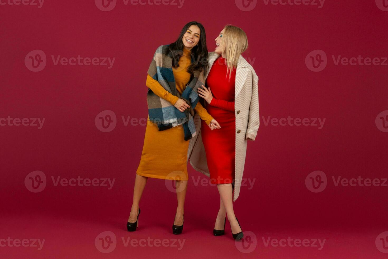 two stylish women in autumn winter fashion dress and coat posing isolated full heigth on red studio background photo