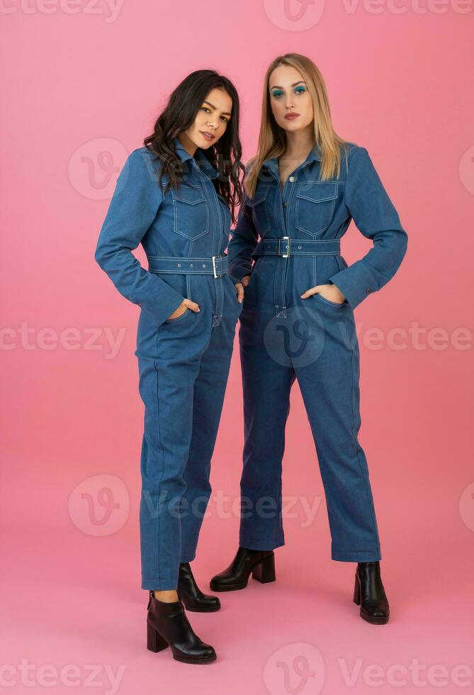 two excited attractive girl friends active women posing on pink background in denim overall having fun together fashion trend photo