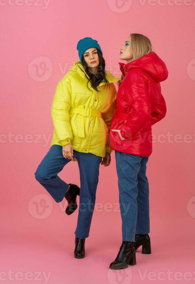 two excited attractive girl friends active women posing on pink background in colorful winter down jacket of bright red and yellow color having fun together, warm coat fashion trend photo