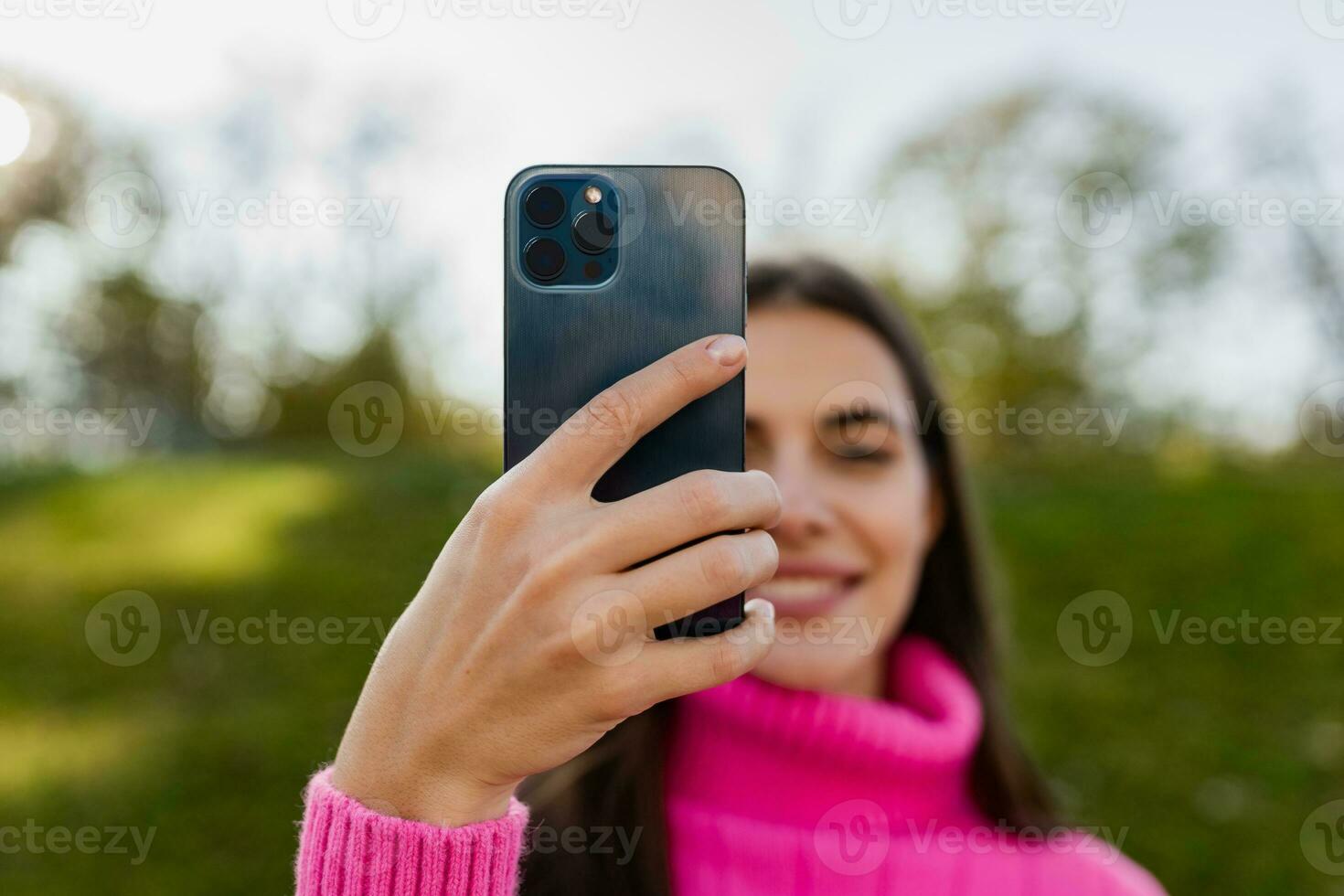 young smiling woman in pink sweater walking in green park using phone photo