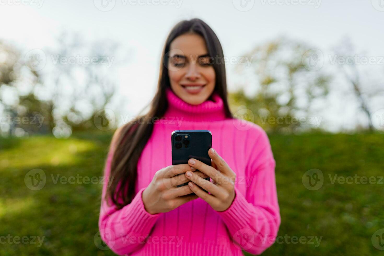 young smiling woman in pink sweater walking in green park using phone photo