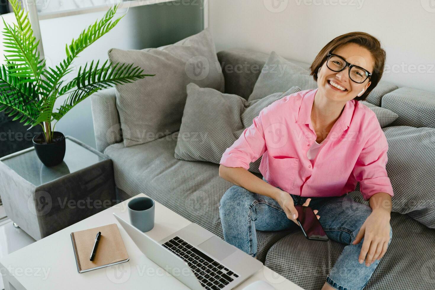 woman in pink shirt sitting relaxed on sofa at home at table working online on laptop photo