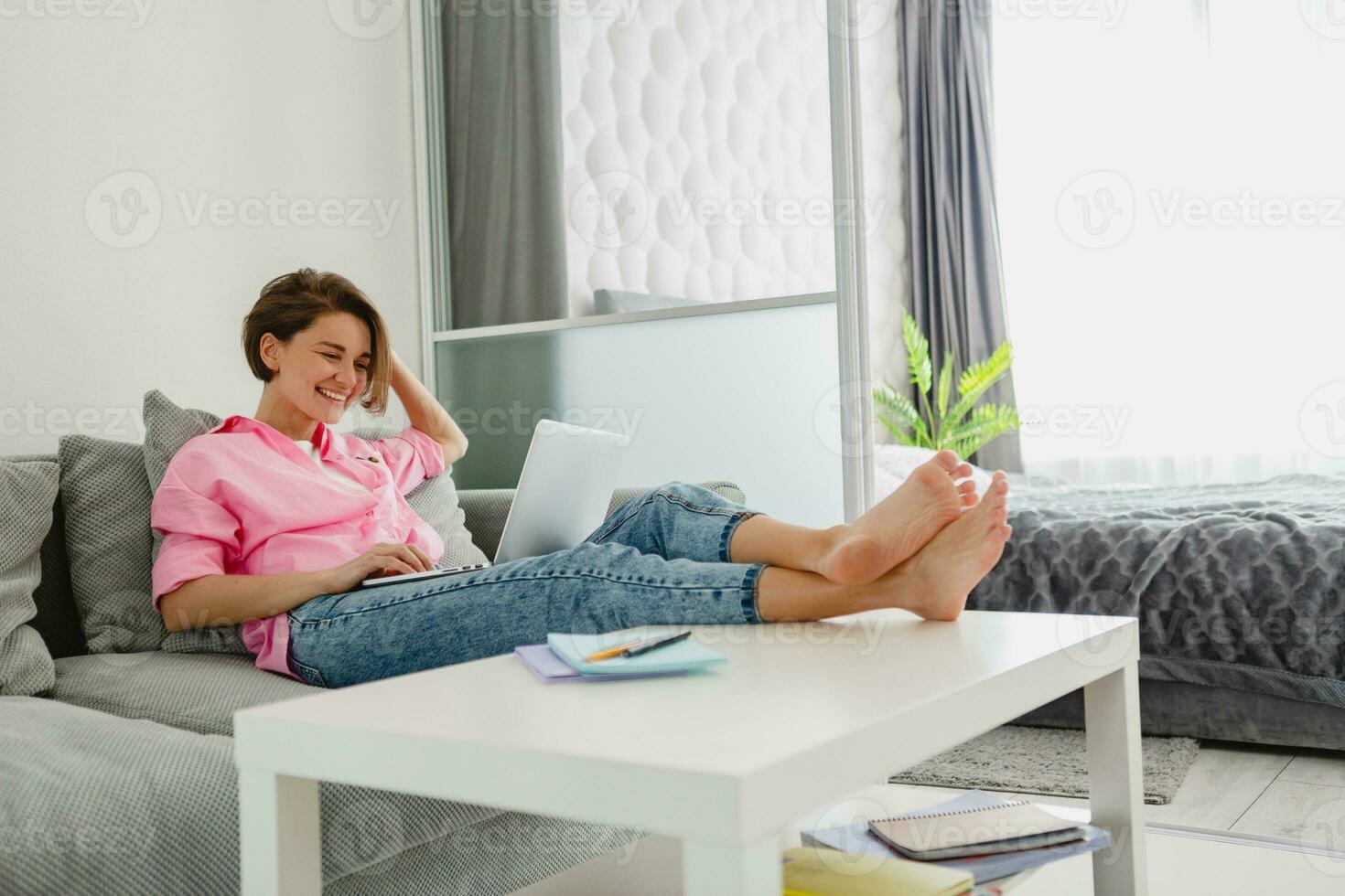 woman in pink shirt sitting relaxed on sofa at home at table working online on laptop photo