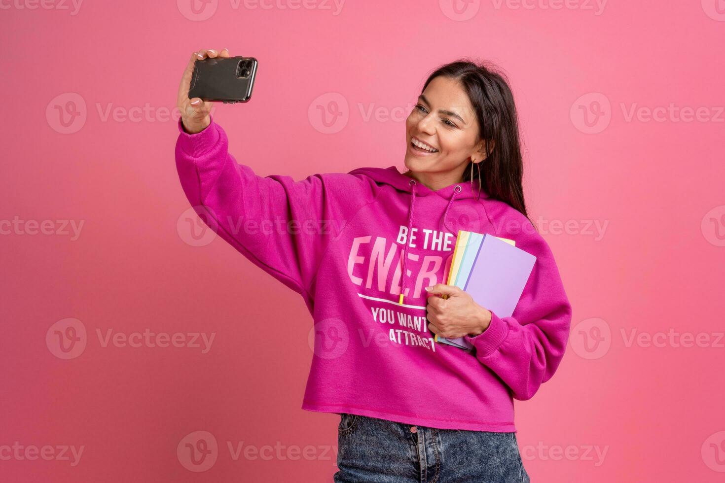 hispanic pretty woman in pink hoodie smiling holding holding notebooks and using smartphone photo