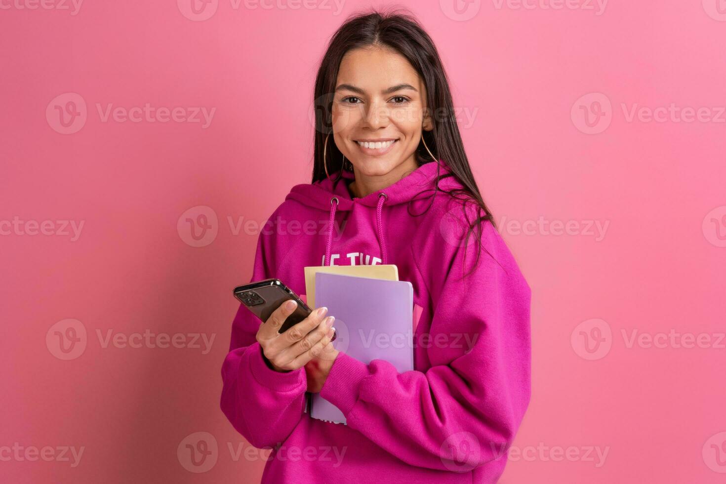 hispanic pretty woman in pink hoodie smiling holding holding notebooks and using smartphone photo