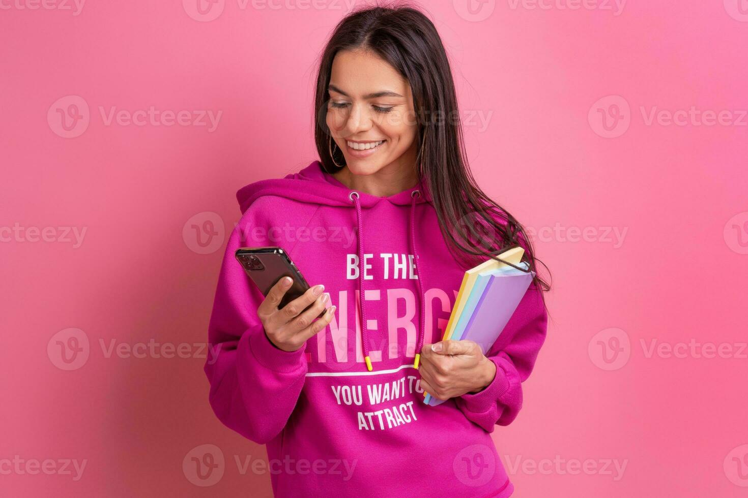 hispanic pretty woman in pink hoodie smiling holding holding notebooks and using smartphone photo