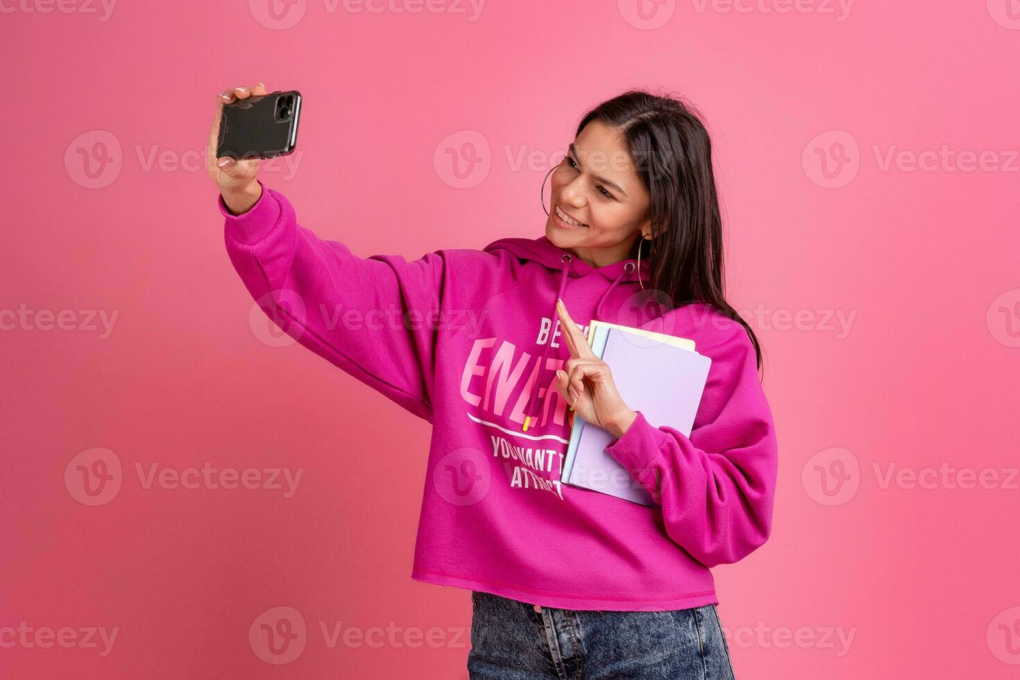 hispanic pretty woman in pink hoodie smiling holding holding notebooks and using smartphone photo