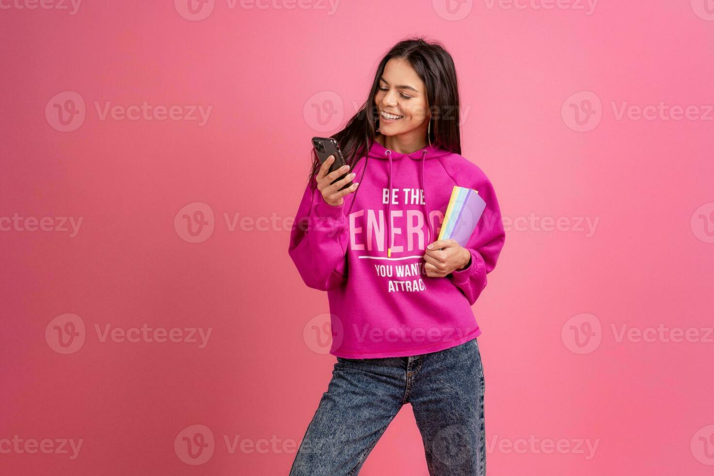 hispanic pretty woman in pink hoodie smiling holding holding notebooks and using smartphone photo