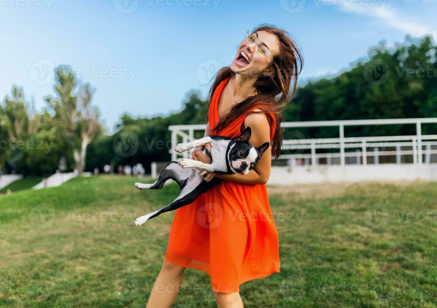young happy smiling woman in orange dress having fun playing with dog in park photo