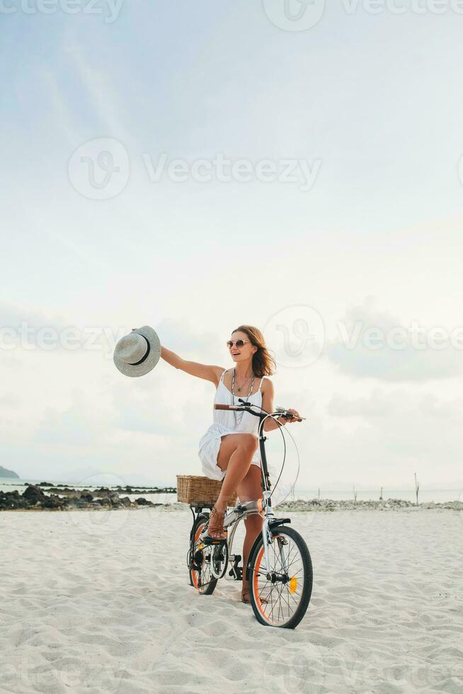 young attractive smiling woman in white dress riding on tropical beach on bicycle photo