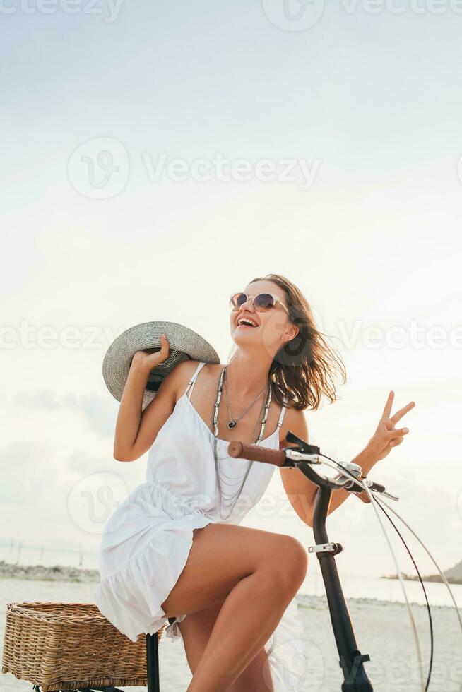 young attractive smiling woman in white dress riding on tropical beach on bicycle photo