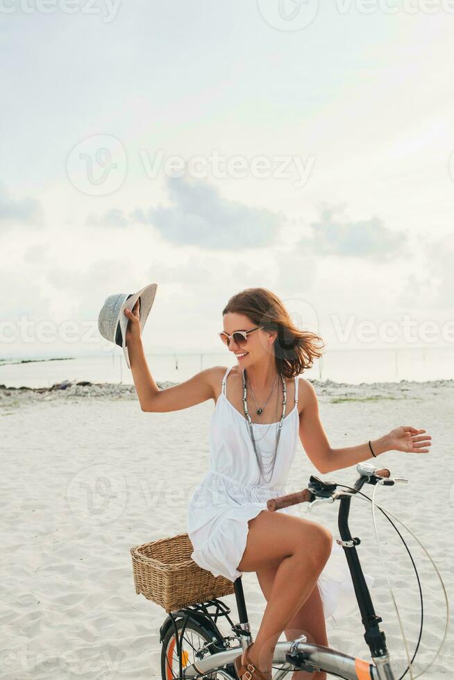 young attractive smiling woman in white dress riding on tropical beach on bicycle photo