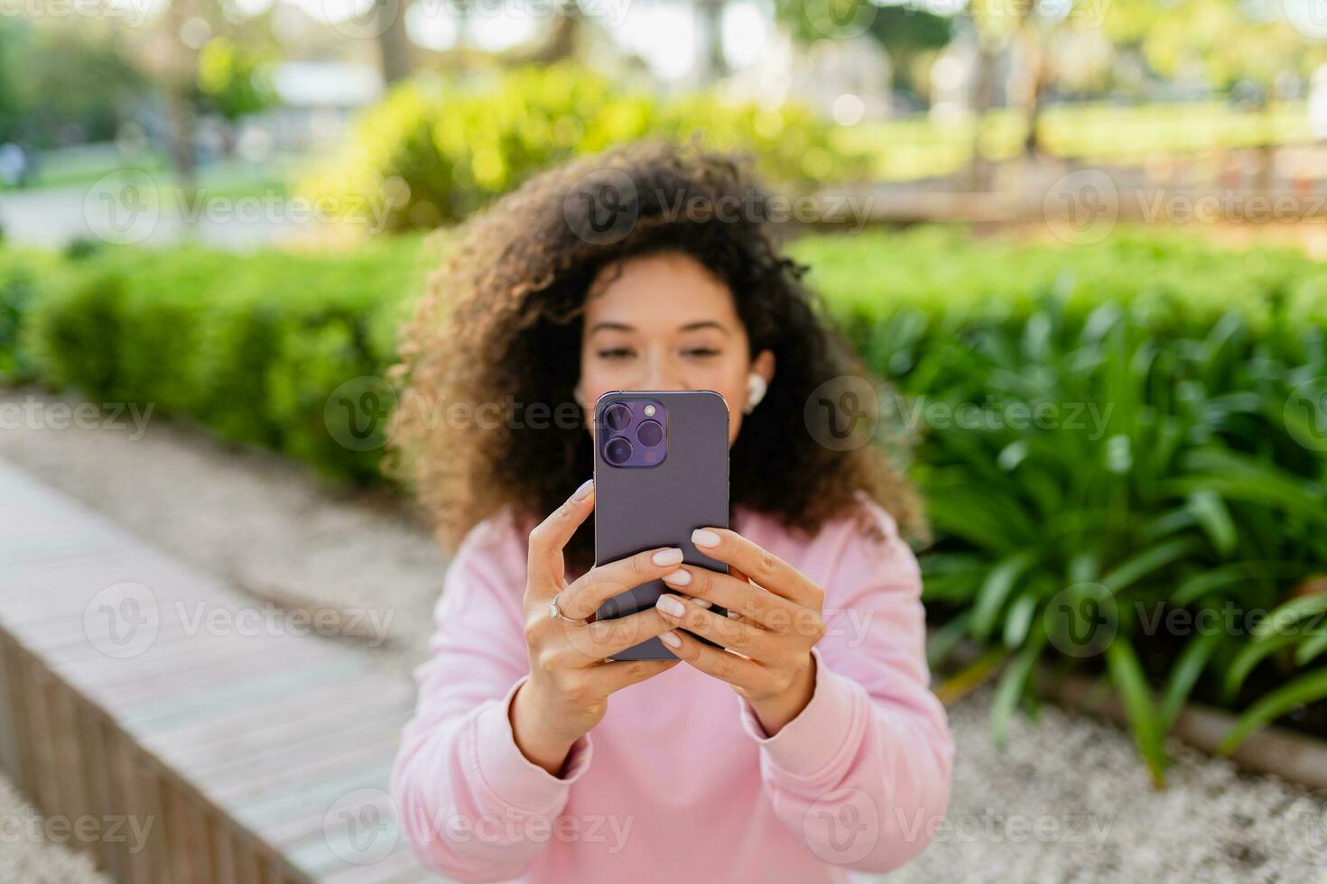 bonito hermosa Rizado joven mujer sentado en parque utilizando smatphone foto