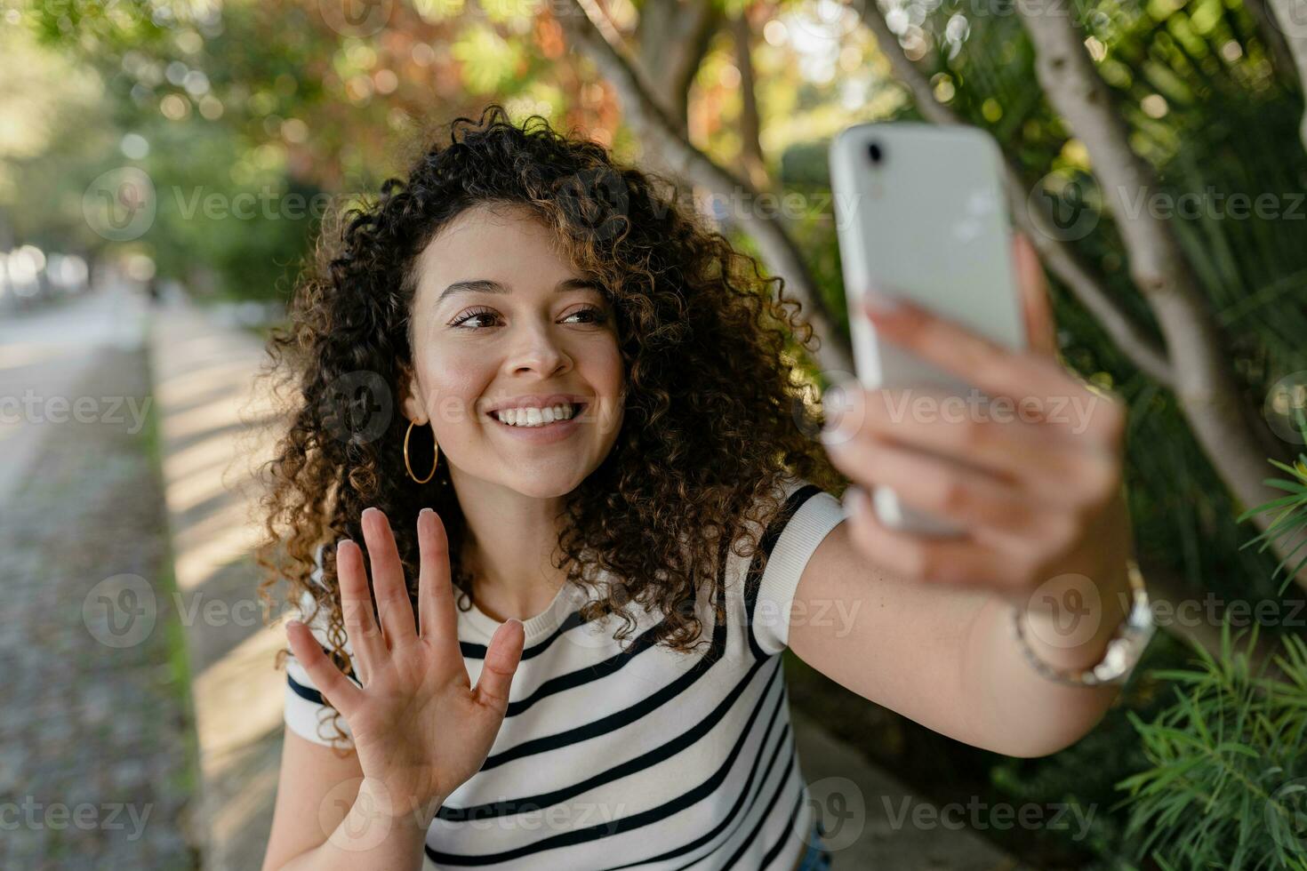 pretty curly woman walking in city street in striped t-shirt, using smart phone photo