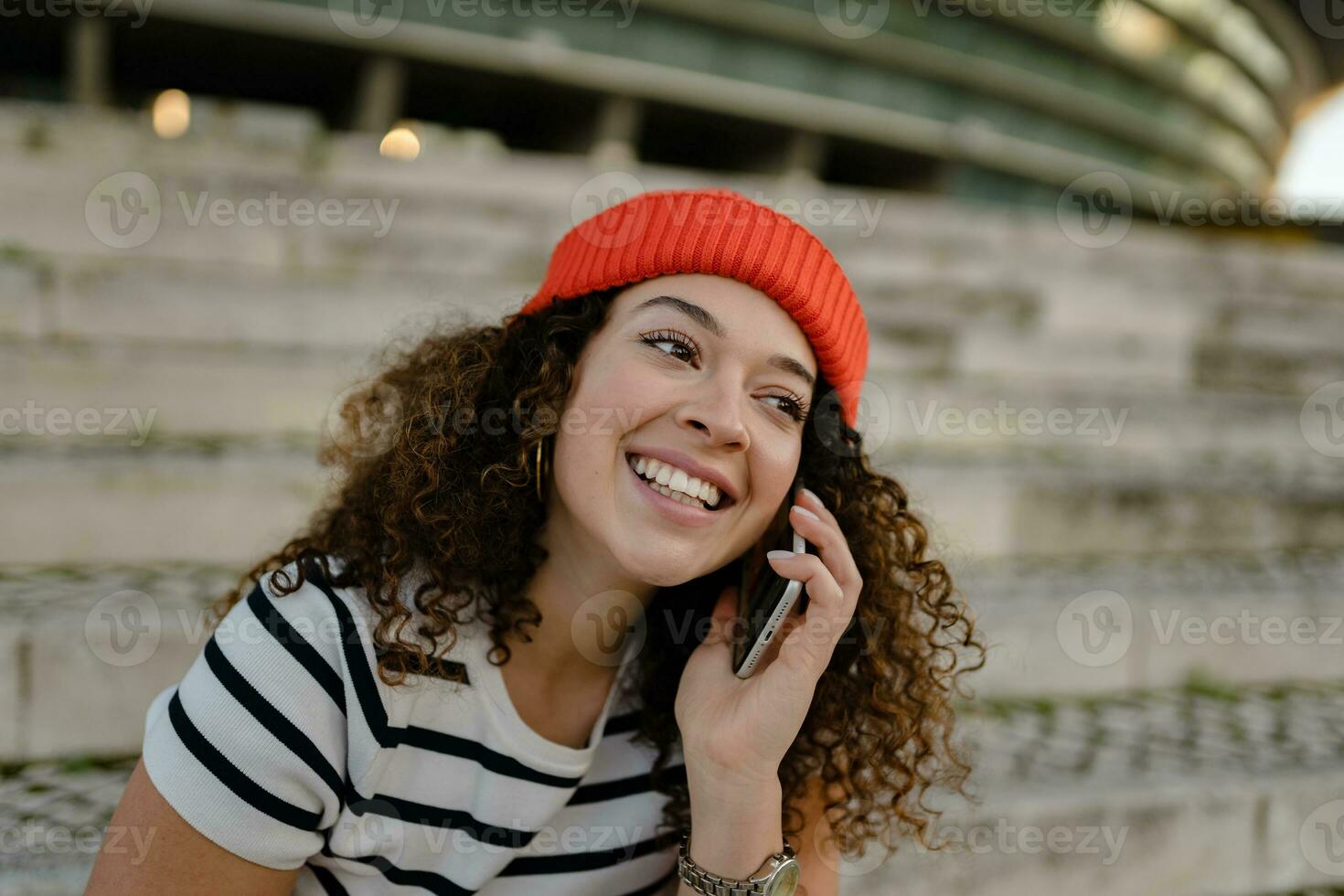 bonito Rizado sonriente mujer sentado en ciudad calle en a rayas camiseta y de punto rojo sombrero, utilizando teléfono inteligente foto