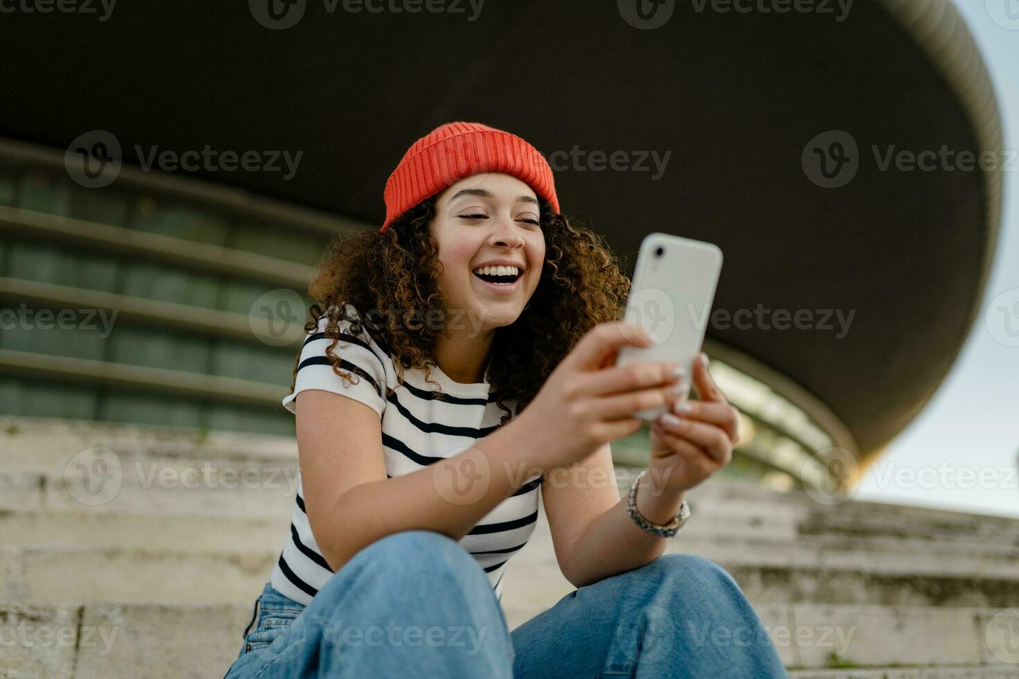 pretty curly smiling woman sitting in city street in striped t-shirt and knitted red hat, using smartphone photo