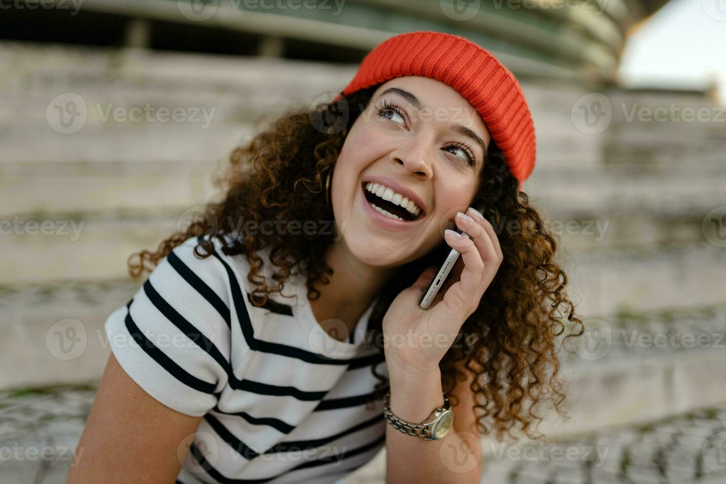 pretty curly smiling woman sitting in city street in striped t-shirt and knitted red hat, using smartphone photo