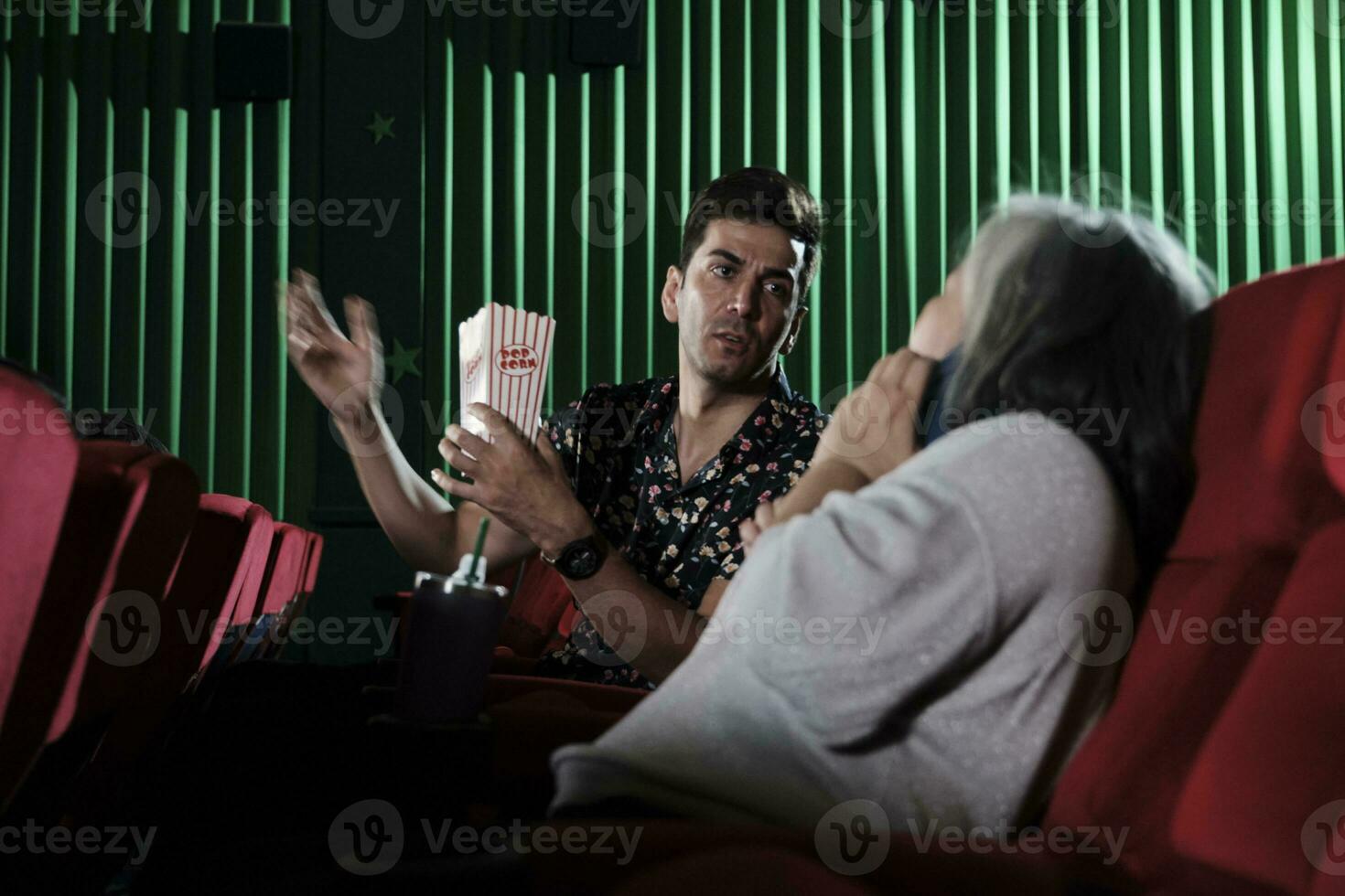 Senior woman using mobile phones in movie theater disturbs male Hispanic and other audiences, bad manners behavior when watching film shows, and are annoyed entertainment lifestyle with smartphone. photo