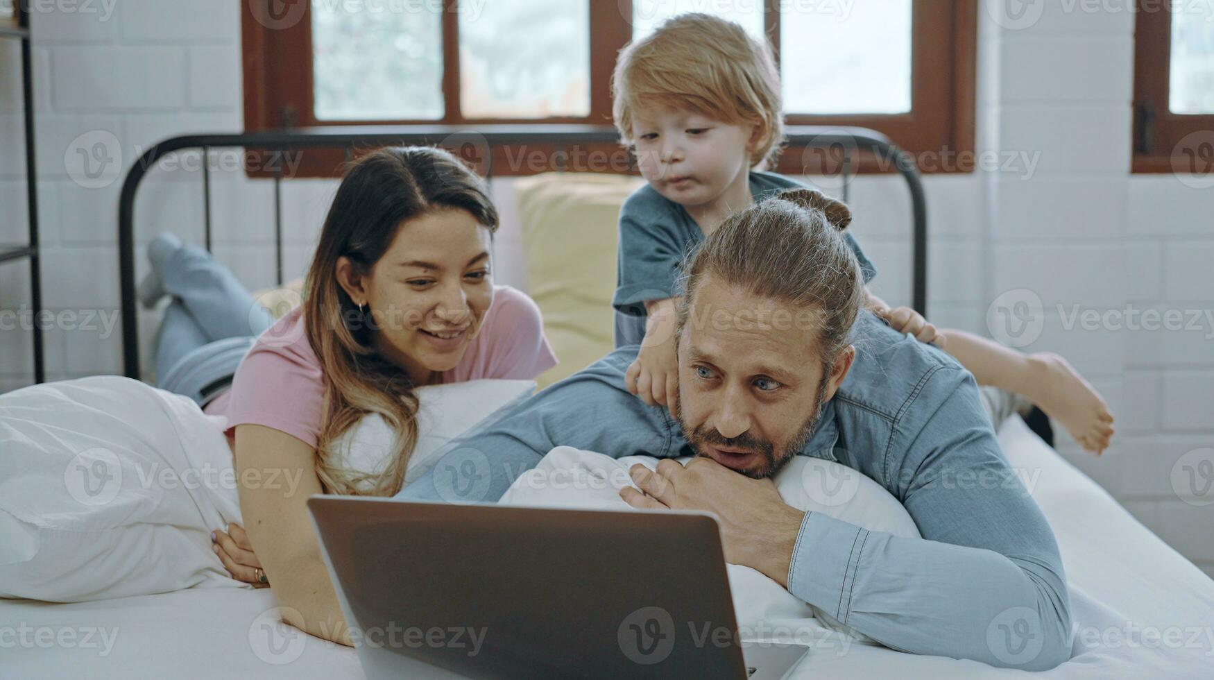 Caucasian family of three using laptop while lying on bed together, browsing internet or watching movie. photo