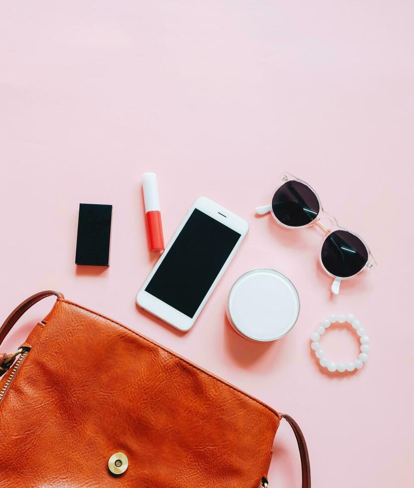 Flat lay of brown leather woman bag open out with cosmetics, accessories and smartphone on pink background photo