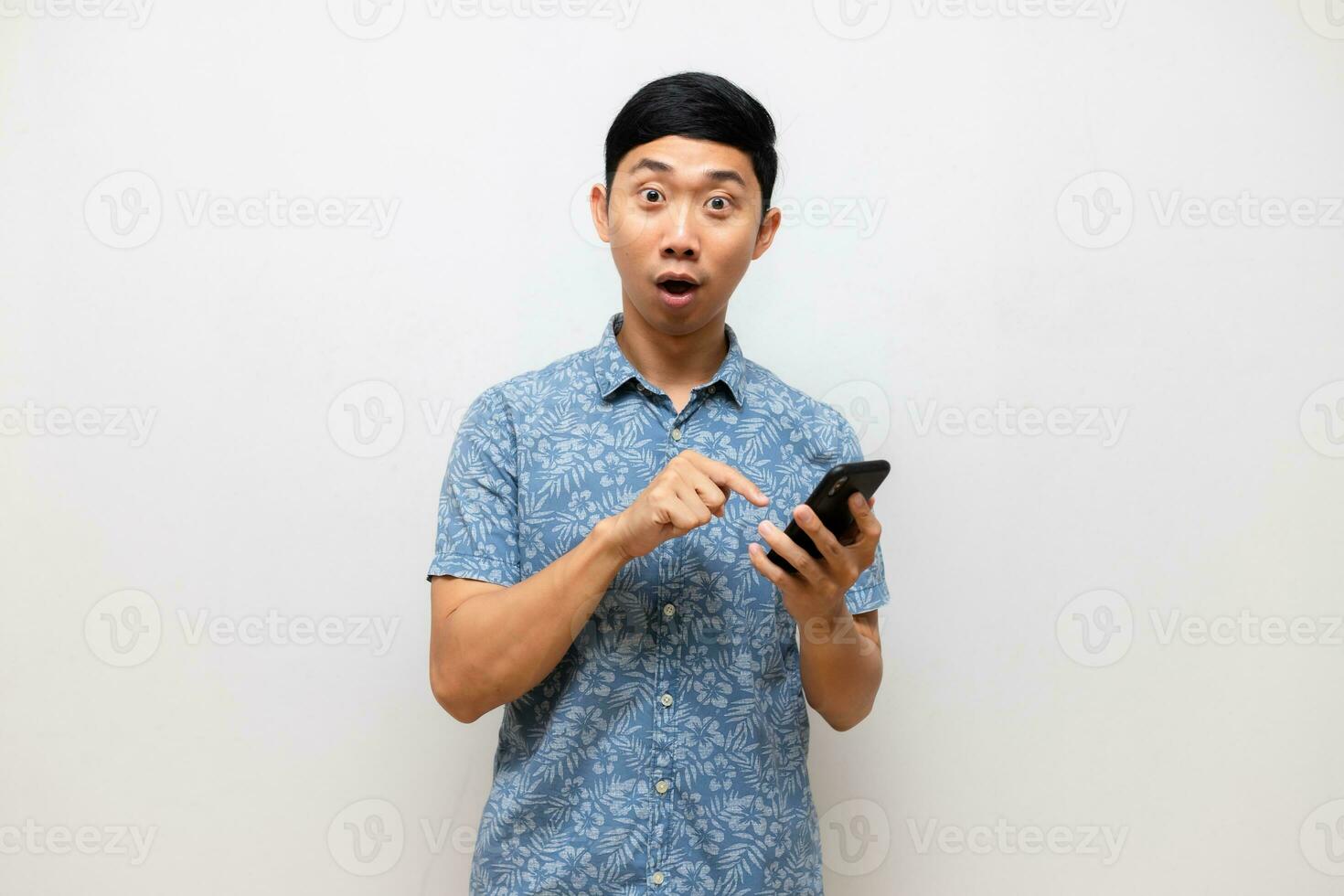 asiático hombre azul camisa siente emocionado y punto dedo a móvil teléfono en mano aislado foto