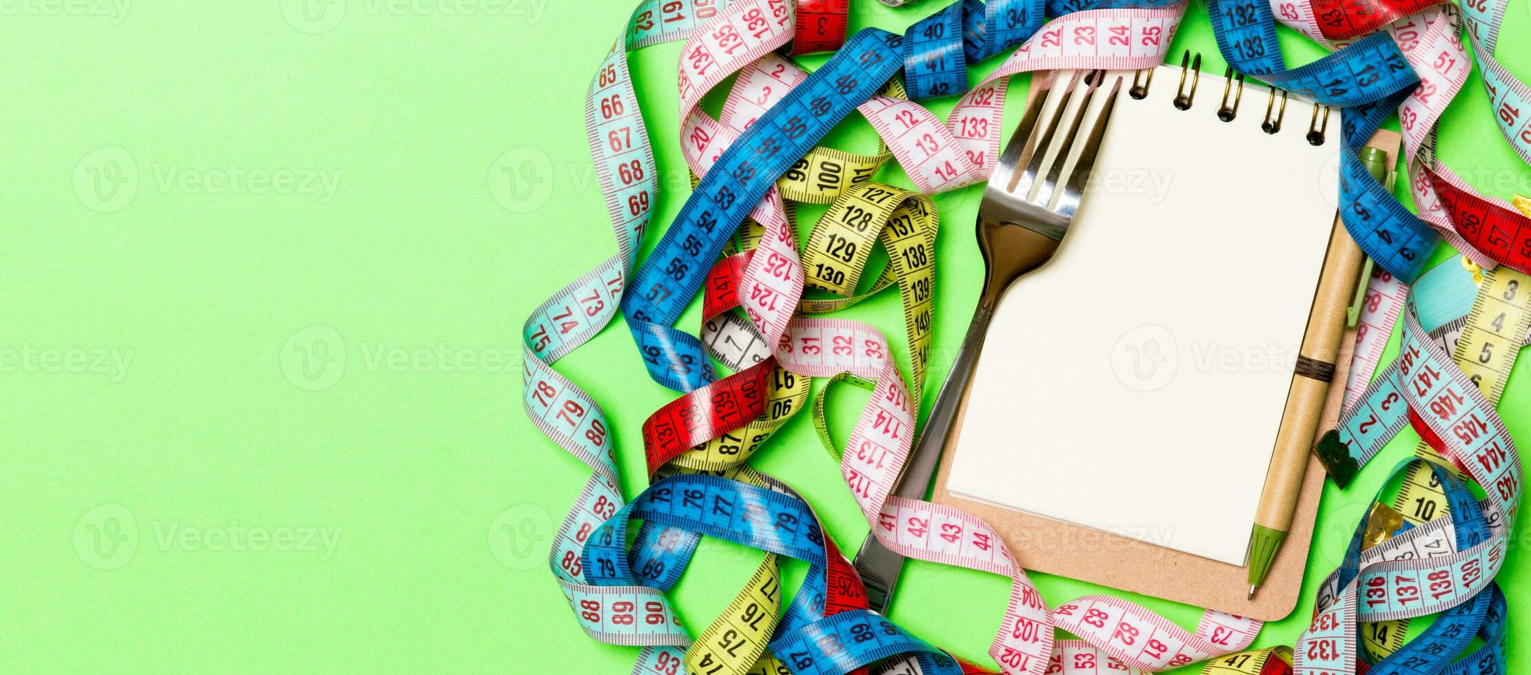 Composition of colorful measuring tapes, fork, pen and notepad on green background. Top view of diet control and empty space for your design photo