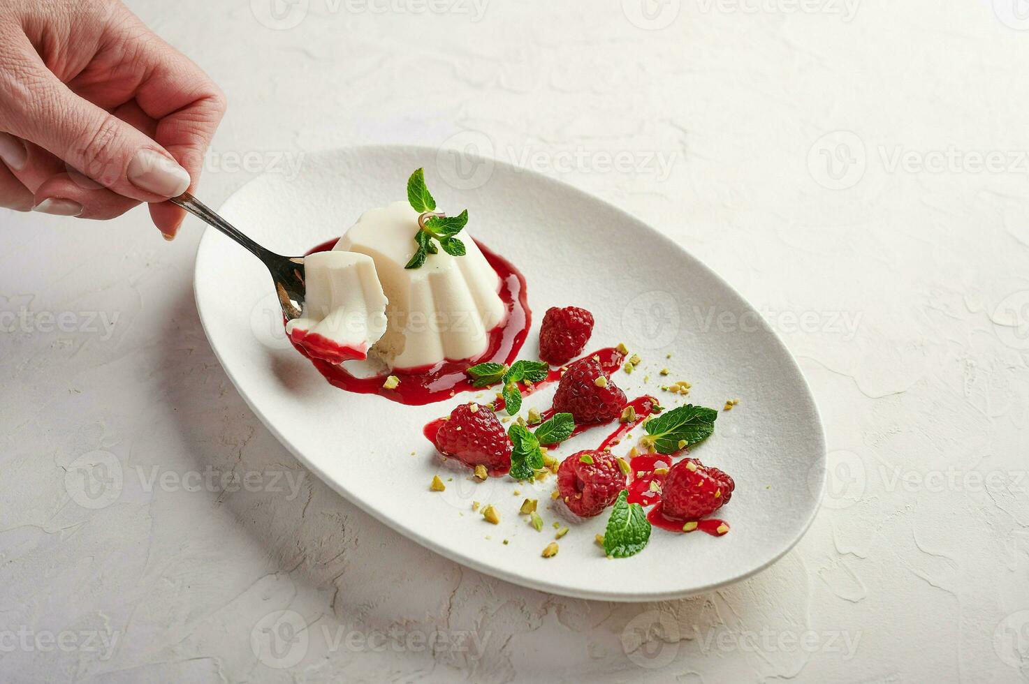 Woman's hand holds a spoon with a piece of panna cotta with syrup, raspberries and mint photo