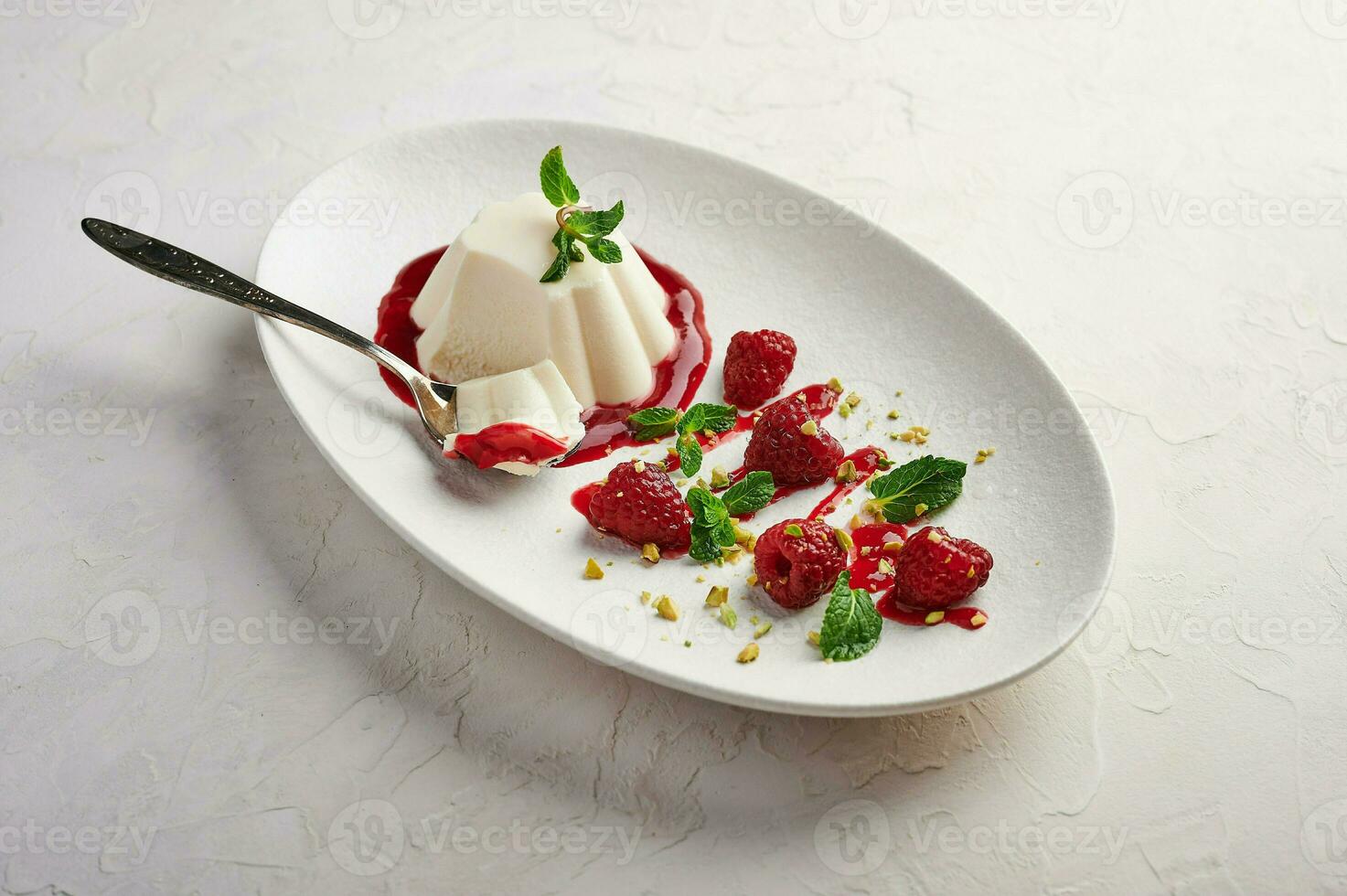 Spoon with a piece of panna cotta with syrup, raspberries and mint in oval plate, food stylind photo