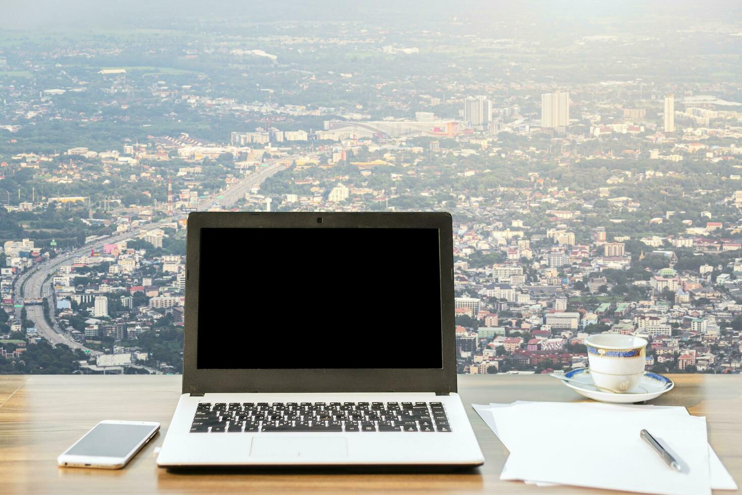Mockup image of laptop with blank black screen,smartphone,coffee cup on wooden table view outdoors of office building dense cityscape high angle view background. photo