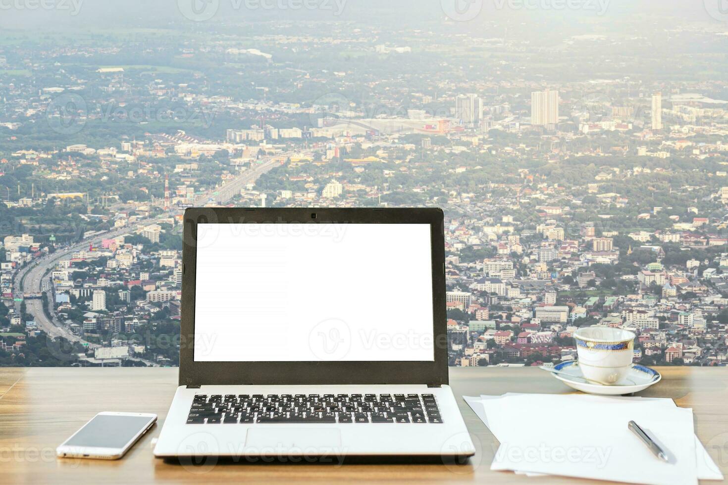 Mockup image of laptop with blank white screen,smartphone,coffee cup on wooden table view outdoors of office building dense cityscape high angle view background. photo