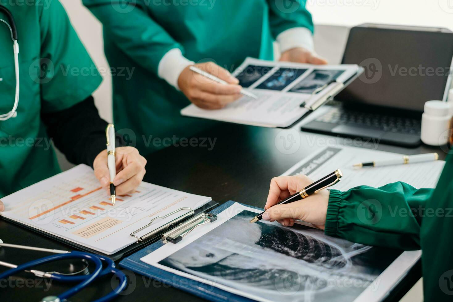 Medical team having a meeting with doctors in white lab coats and surgical scrubs seated at a table discussing a patients working online using computers in the medical photo