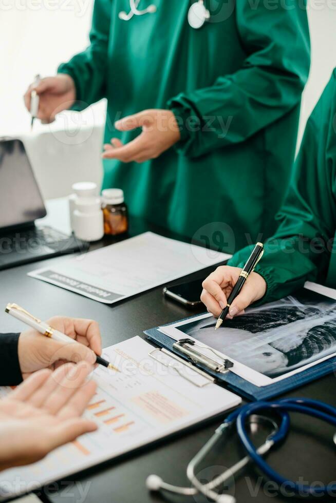 Medical team having a meeting with doctors in white lab coats and surgical scrubs seated at a table discussing a patients working online using computers in the medical photo