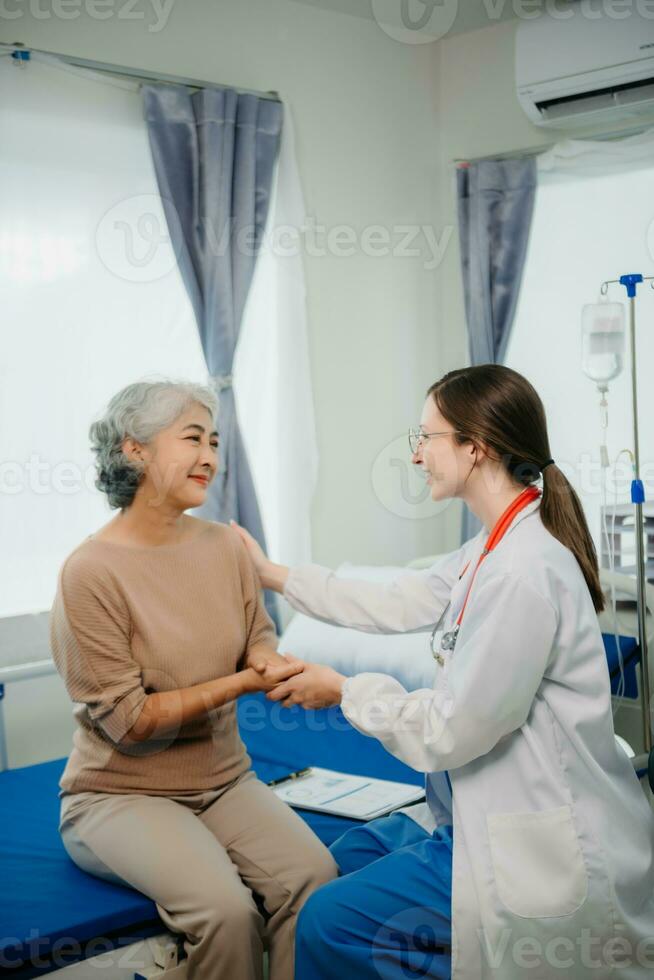Doctor in white suit take notes while discussing and Asian elderly, woman patient who lying on bed with receiving saline solution in hospital or clinic. photo