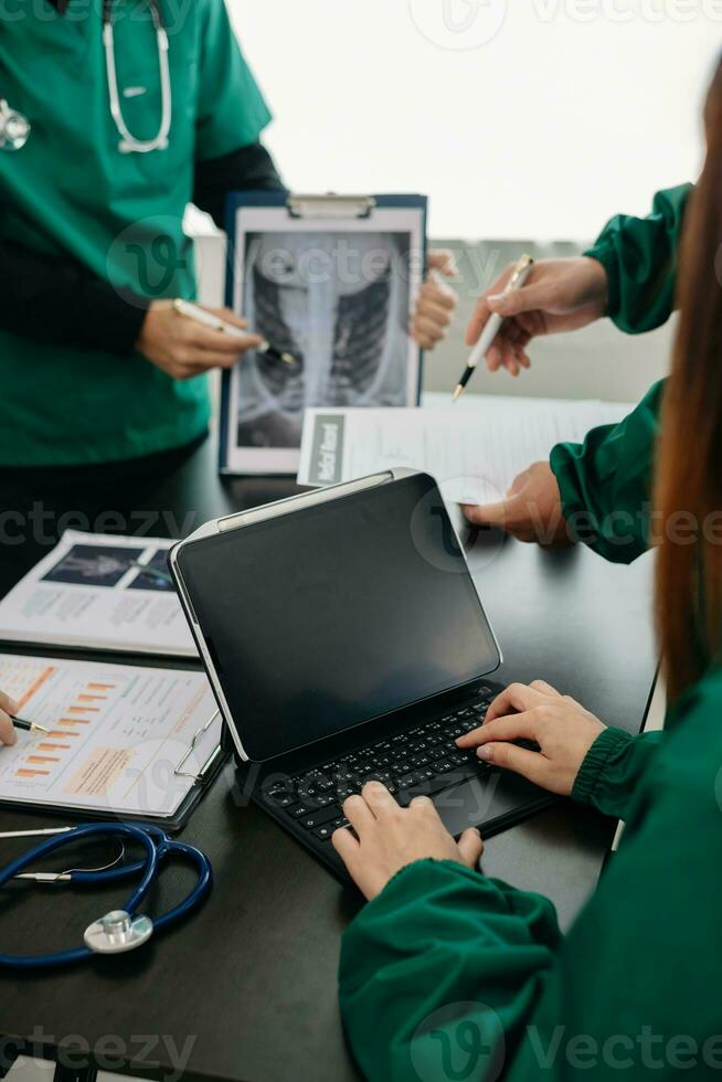 Medical team having a meeting with doctors in white lab coats and surgical scrubs seated at a table discussing a patients working online using computers in the medical photo
