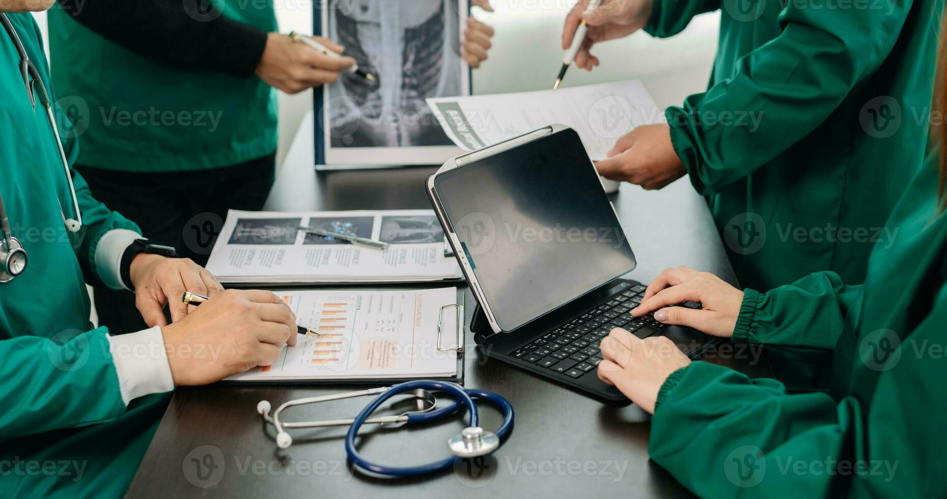 Medical team having a meeting with doctors in white lab coats and surgical scrubs seated at a table discussing a patients working online using computers in the medical photo
