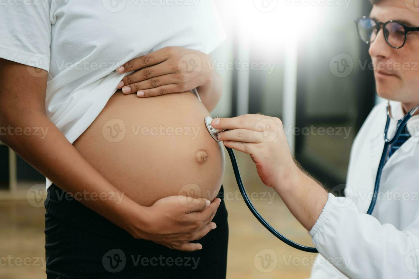 Pregnant african woman has appointment with doctor at clinic. Male gynaecologist OB GYN medic specialist with stethoscope listens to baby's heartbeat in mother's belly. photo