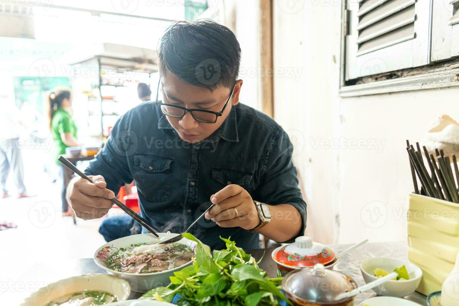A man using chopsticks and spoon eating traditional Pho Bo vietnamese soup with beef and rice noodles on a metal table, real scene in local restaurant photo