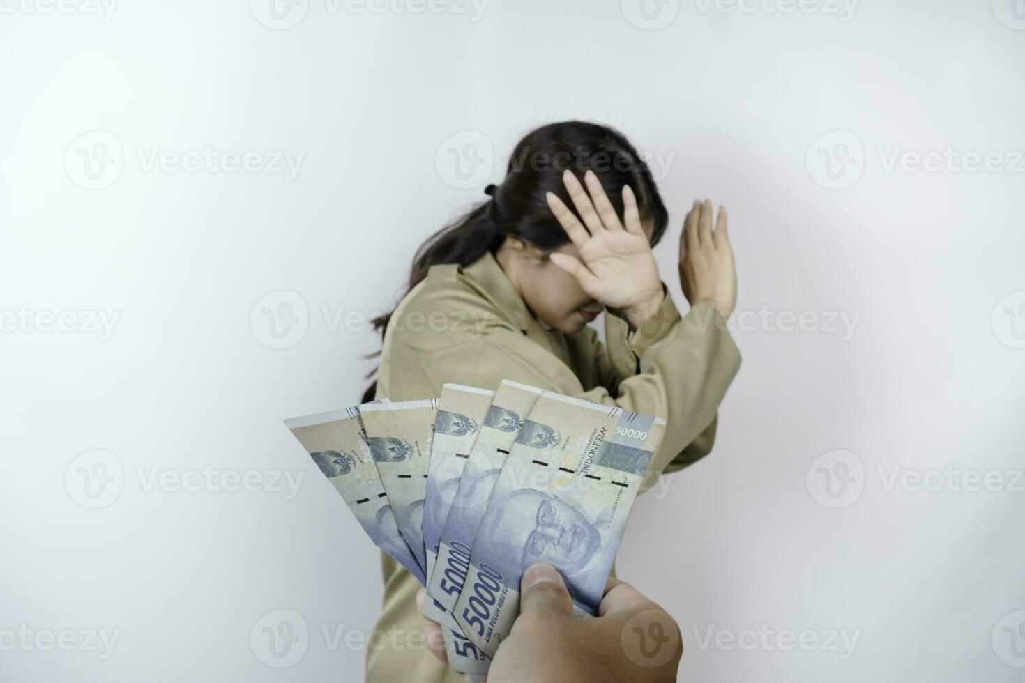 A serious young government worker woman is wearing khaki uniform gesturing rejection to cash money in Indonesian rupiah isolated by white background photo