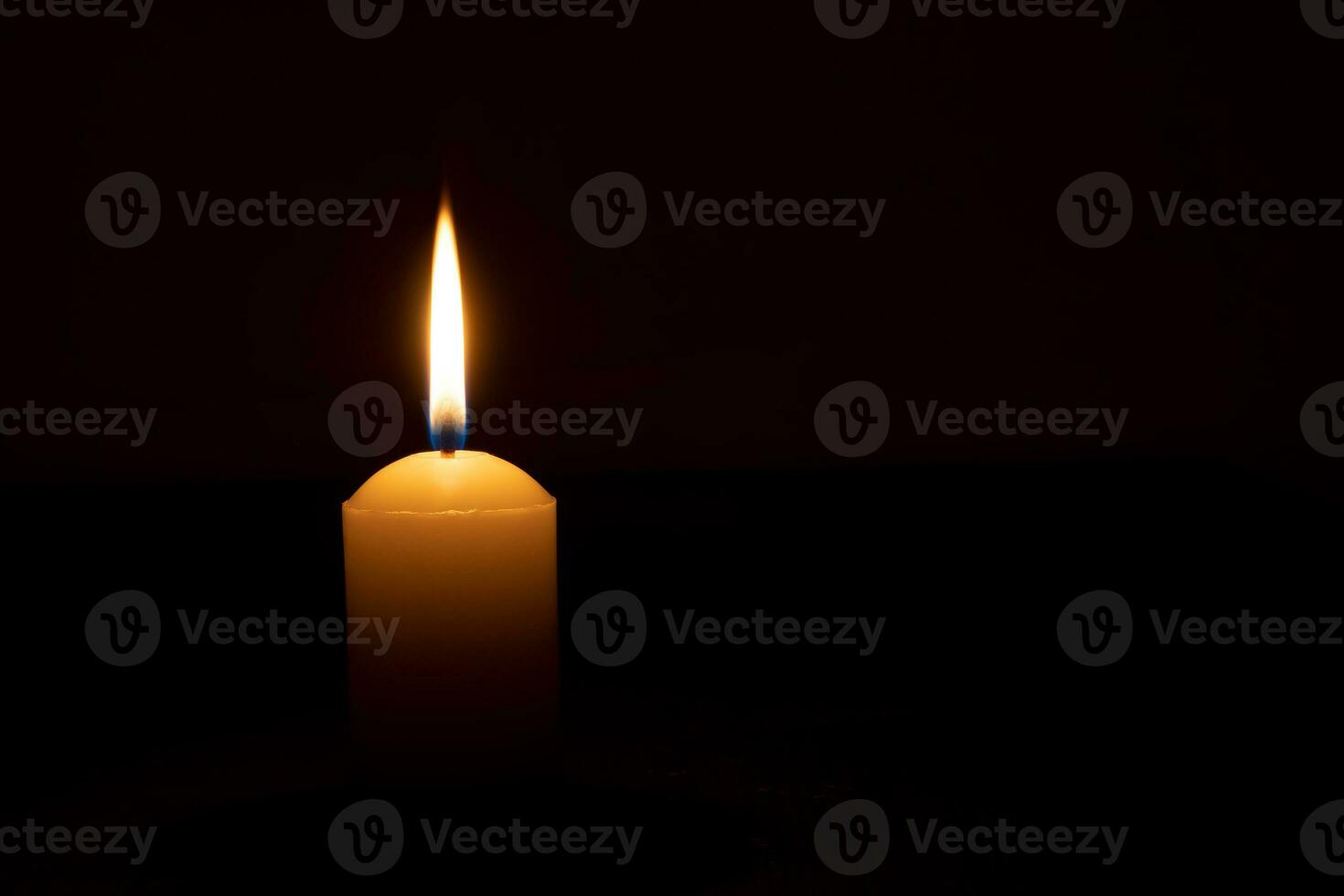 Single burning candle flame or light glowing on a big white candle on black or dark background on table in church for Christmas, funeral or memorial service with copy space. photo