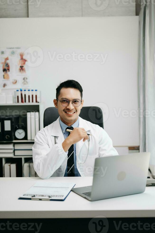 médico médico sentar en mesa en hospital. atractivo terapeuta masculino facultativo farmacéutico sonrisa y utilizar tableta y ordenador portátil. después éxito trabajo desde tratamiento foto