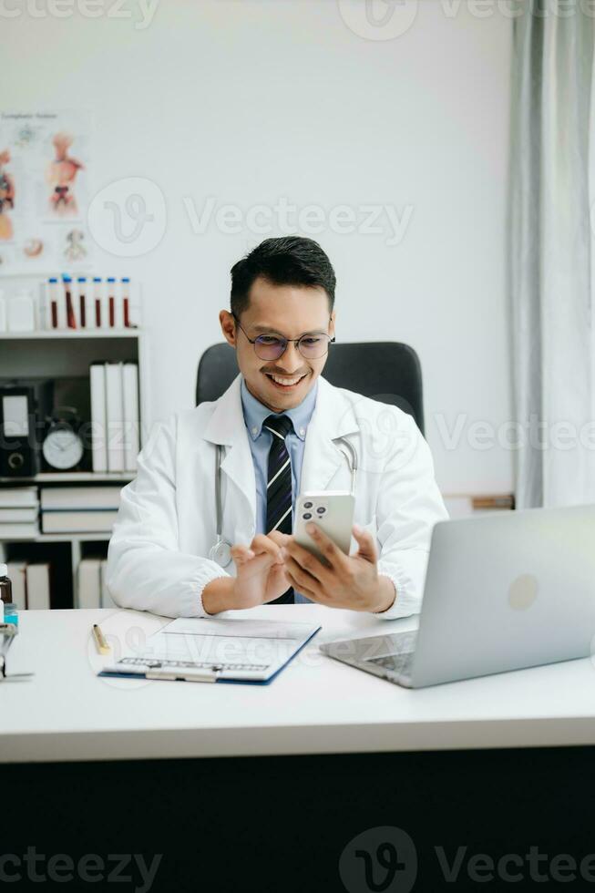 médico médico sentar en mesa en hospital. atractivo terapeuta masculino facultativo farmacéutico sonrisa y utilizar tableta y ordenador portátil. después éxito trabajo desde tratamiento foto